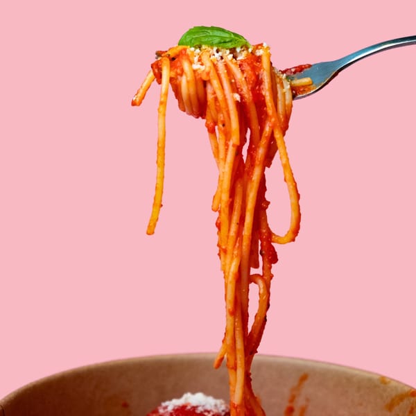 Spaghetti with red sauce, parmesan, and basil on a fork, lifted from a bowl against a pink background.