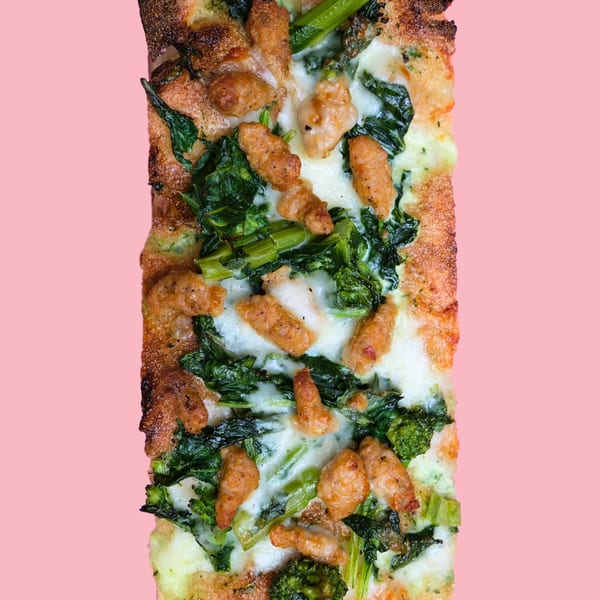 Rectangular pizza with sausage, broccoli rabe, and melted cheese on a pink background.