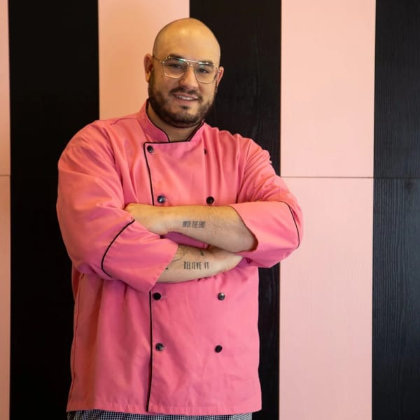 Smiling Pizza Collective chef in a pink uniform with crossed arms against a pink and black striped wall.