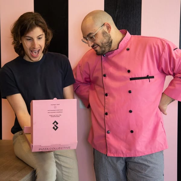 A chef in a pink uniform and a customer excitedly open a Pizza Collective pizza box.