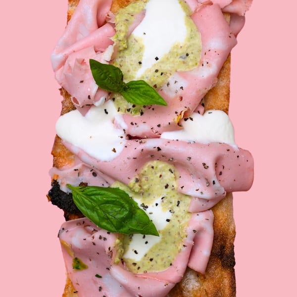 Mortadella, burrata, pistachio pesto, and basil on focaccia bread against a pink background.