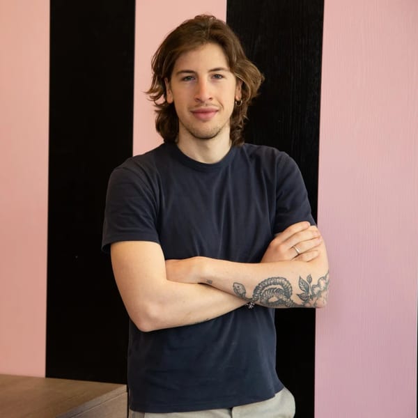 A young man with long brown hair and tattoos on his arms stands against a pink and black striped wall.